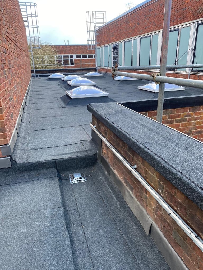 Industrial flat rooftop with dark membrane surface, multiple skylights, brick walls, and metal railings