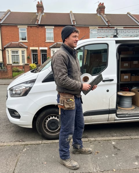 A tradesman in a black beanie and brown jacket stands beside a white work van on a residential street, holding a tablet or clipboard