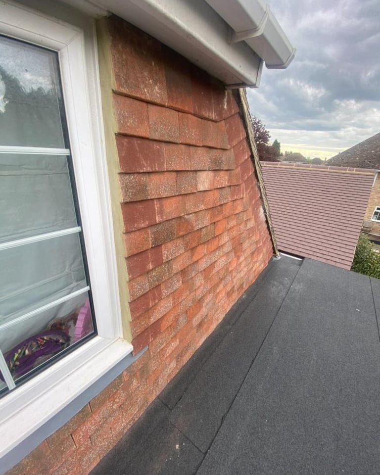 Exterior view of a house roofline showing red brick wall, white window frame, and gray tiled roof against a cloudy sky