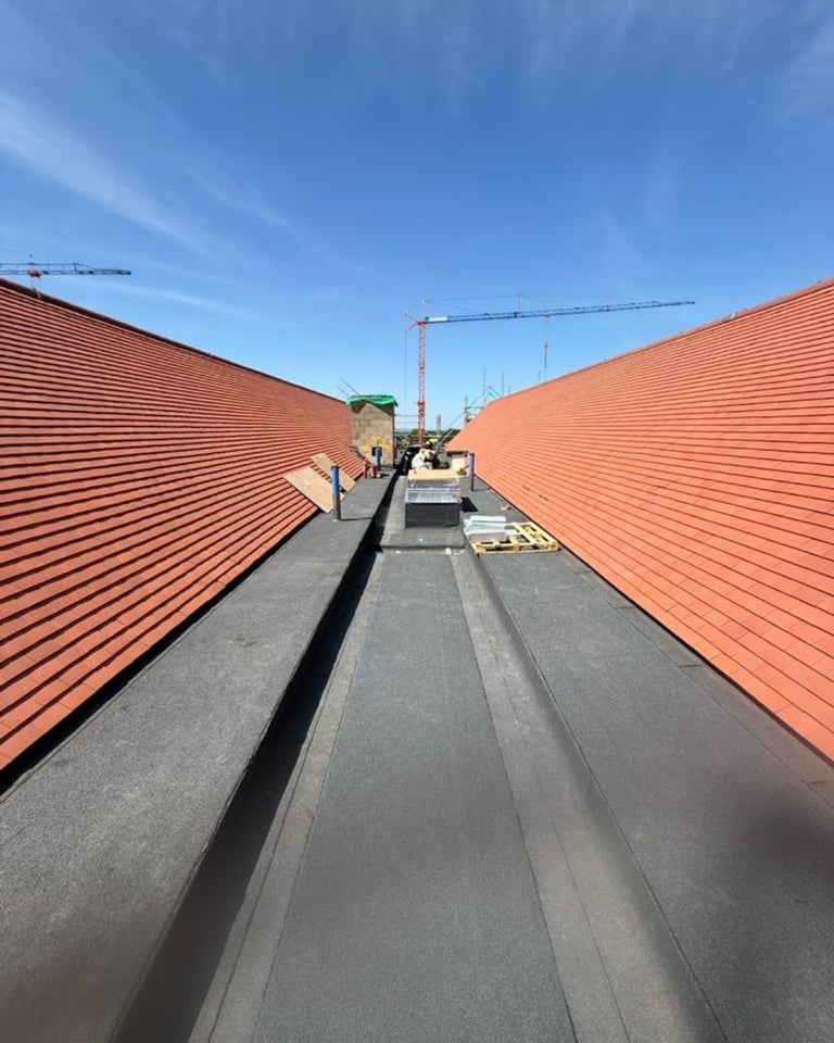 Aerial view of a construction site rooftop with red metal corrugated panels, dark walkway, cranes, and clear blue sky